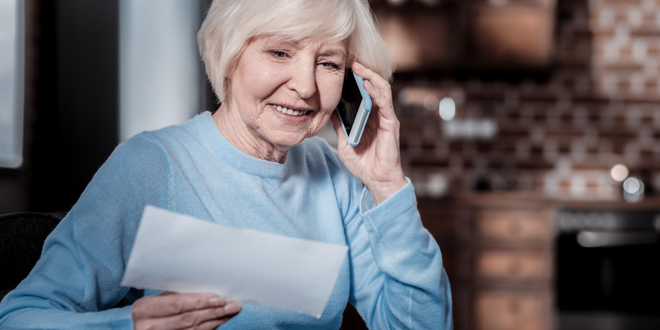 Eine Seniorin telefoniert gut gelaunt mit einem Smartphone am Ohr. Sie hält ein Dokument in der rechten Hand.