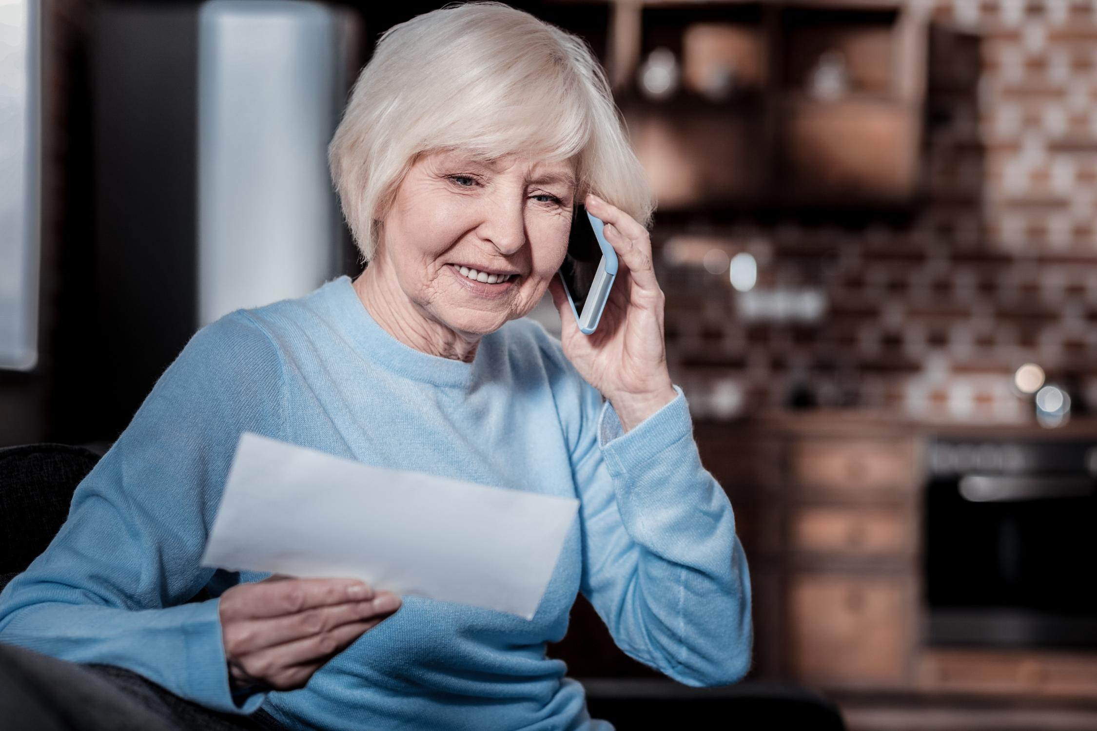 Eine Seniorin telefoniert gut gelaunt mit einem Smartphone am Ohr. Sie hält ein Dokument in der rechten Hand.