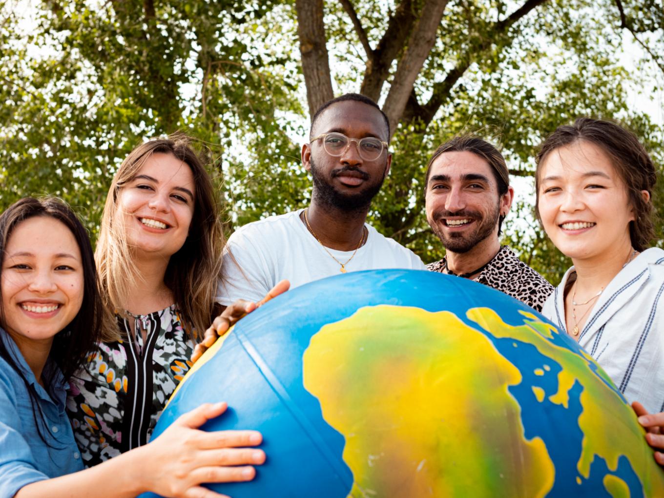 Junge Erwachsene unterschiedlicher Herkunft halten gemeinsam einen aufblasbaren Globus. Sie stehen in der Natur vor einem großen Baum.