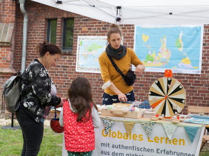 Eine Frau steht hinter einem Stand: Globales Lernen. Davor steht eine Mutter mit Kind