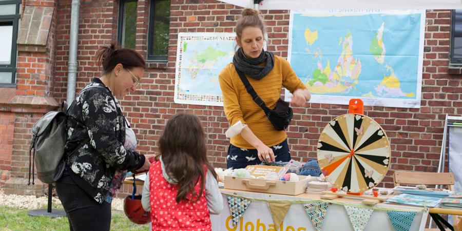 Eine Frau steht hinter einem Stand: Globales Lernen. Davor steht eine Mutter mit Kind