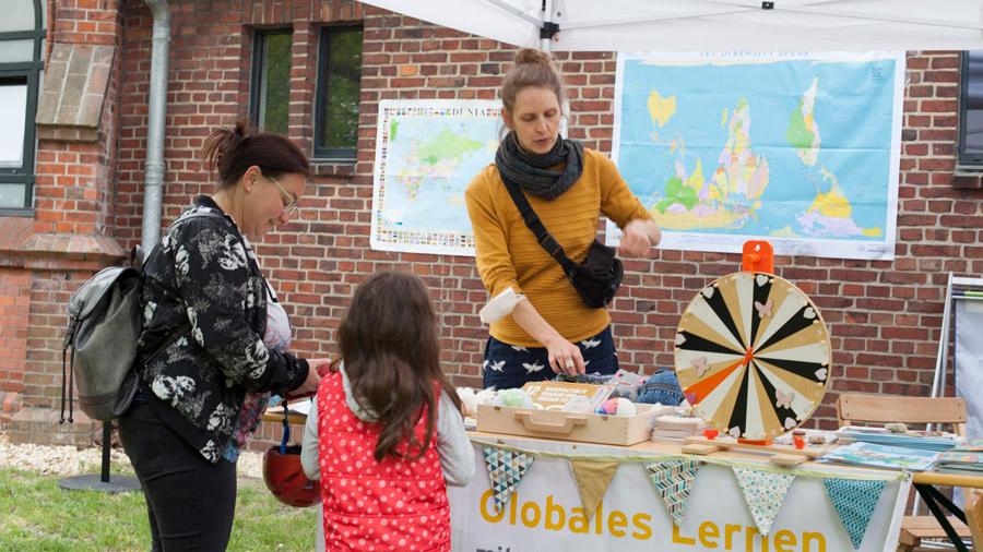 Eine Frau steht hinter einem Stand: Globales Lernen. Davor steht eine Mutter mit Kind