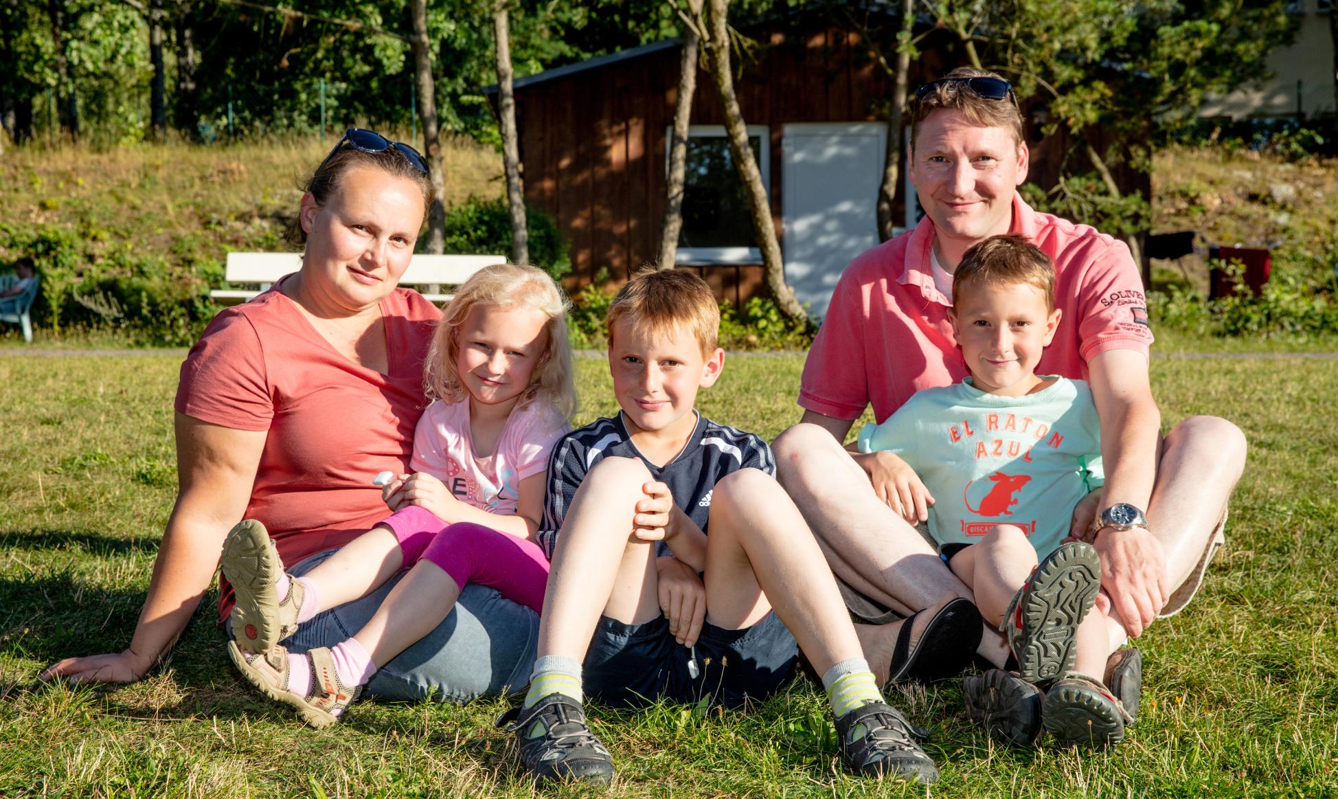 Vater, Mutter, zwei Söhne und eine Tochter sitzen gemeinsam und entspannt auf einer Wiese und schauen lächelnd in die Kamera