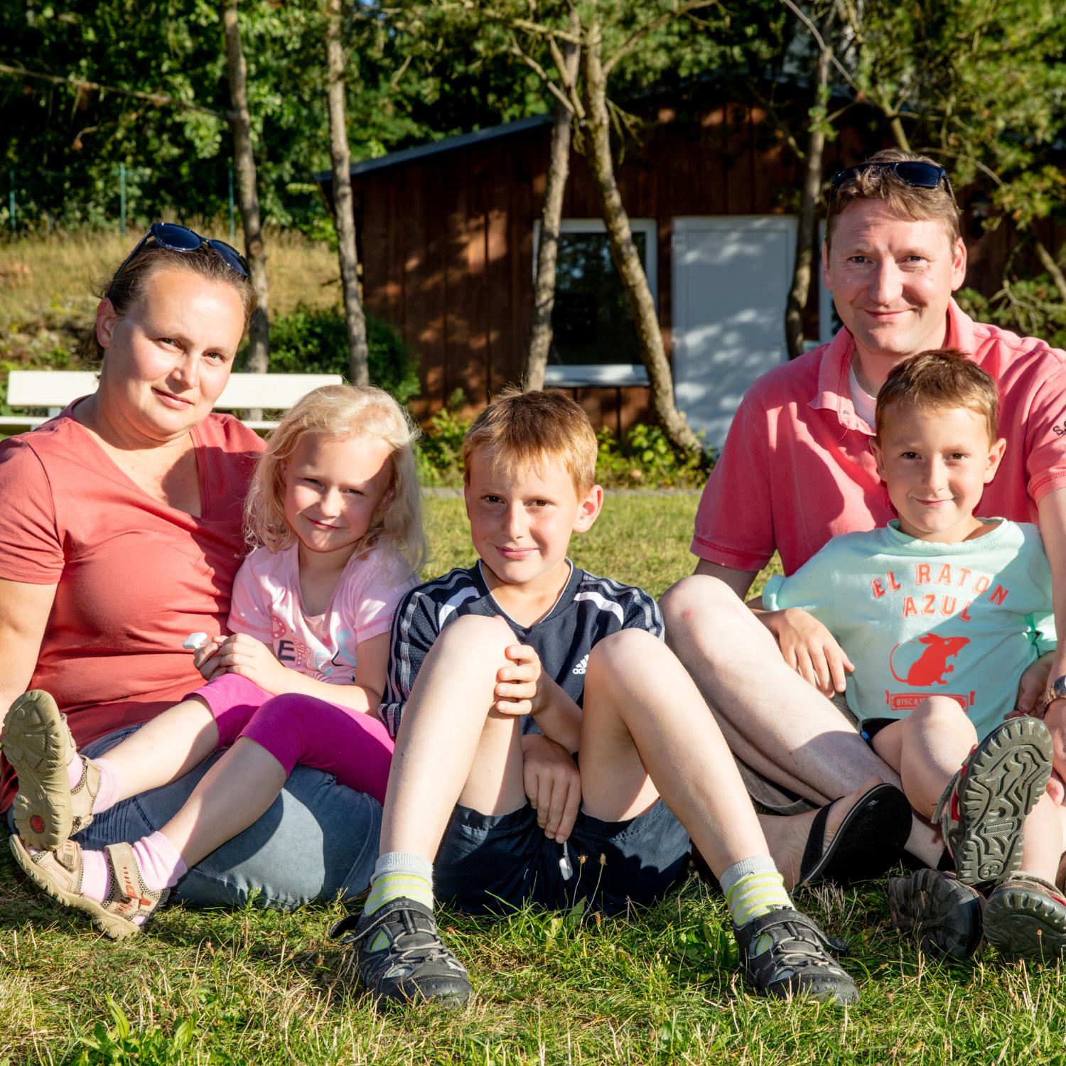 Vater, Mutter, zwei Söhne und eine Tochter sitzen gemeinsam und entspannt auf einer Wiese und schauen lächelnd in die Kamera