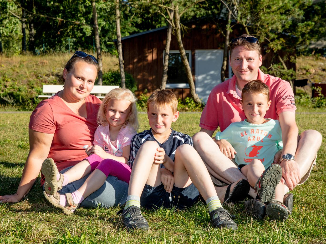 Vater, Mutter, zwei Söhne und eine Tochter sitzen gemeinsam und entspannt auf einer Wiese und schauen lächelnd in die Kamera
