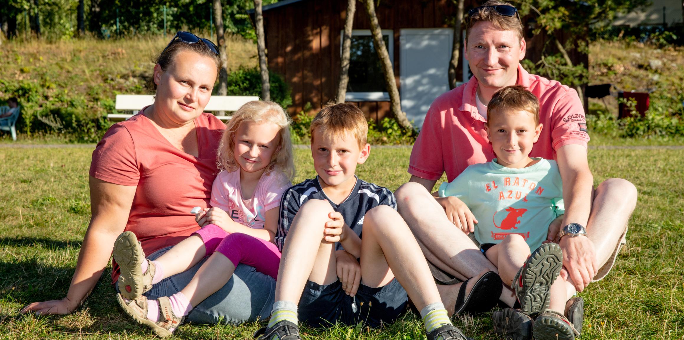 Vater, Mutter, zwei Söhne und eine Tochter sitzen gemeinsam und entspannt auf einer Wiese und schauen lächelnd in die Kamera