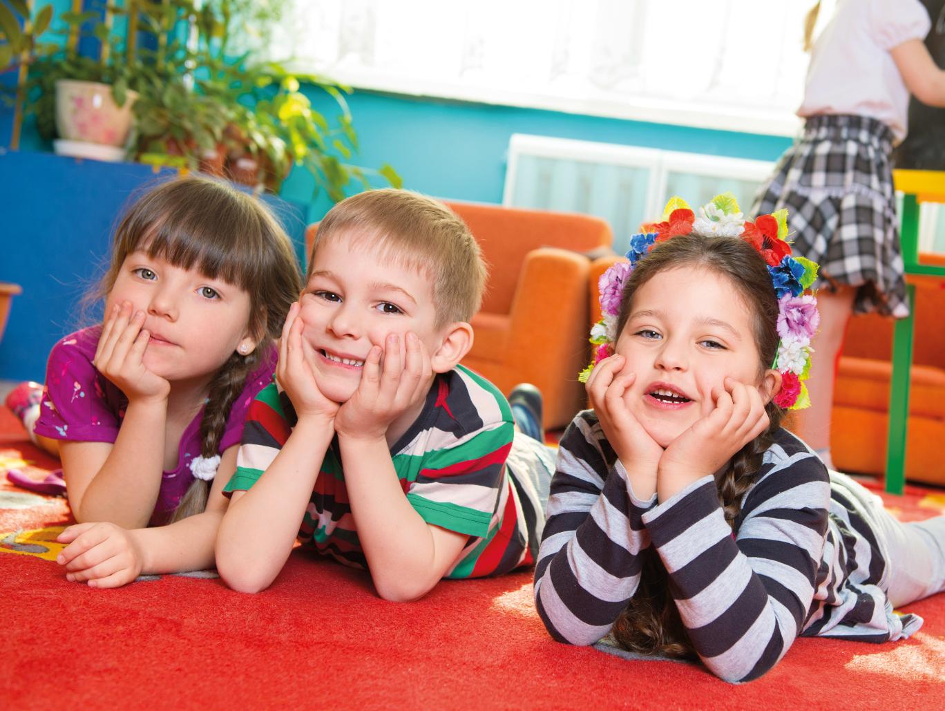 Ein Junge und zwei Mädchen schauen im Kindergarten lachend in die Kamera. Sie liegen auf einer Spielmatte und haben Ihre Köpfe in die Hände gestützt