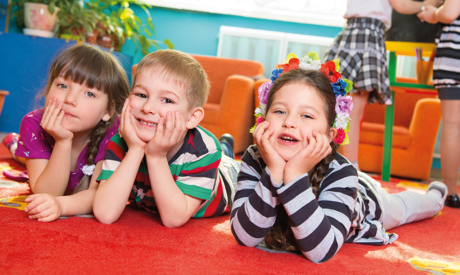 Ein Junge und zwei Mädchen schauen im Kindergarten lachend in die Kamera. Sie liegen auf einer Spielmatte und haben Ihre Köpfe in die Hände gestützt