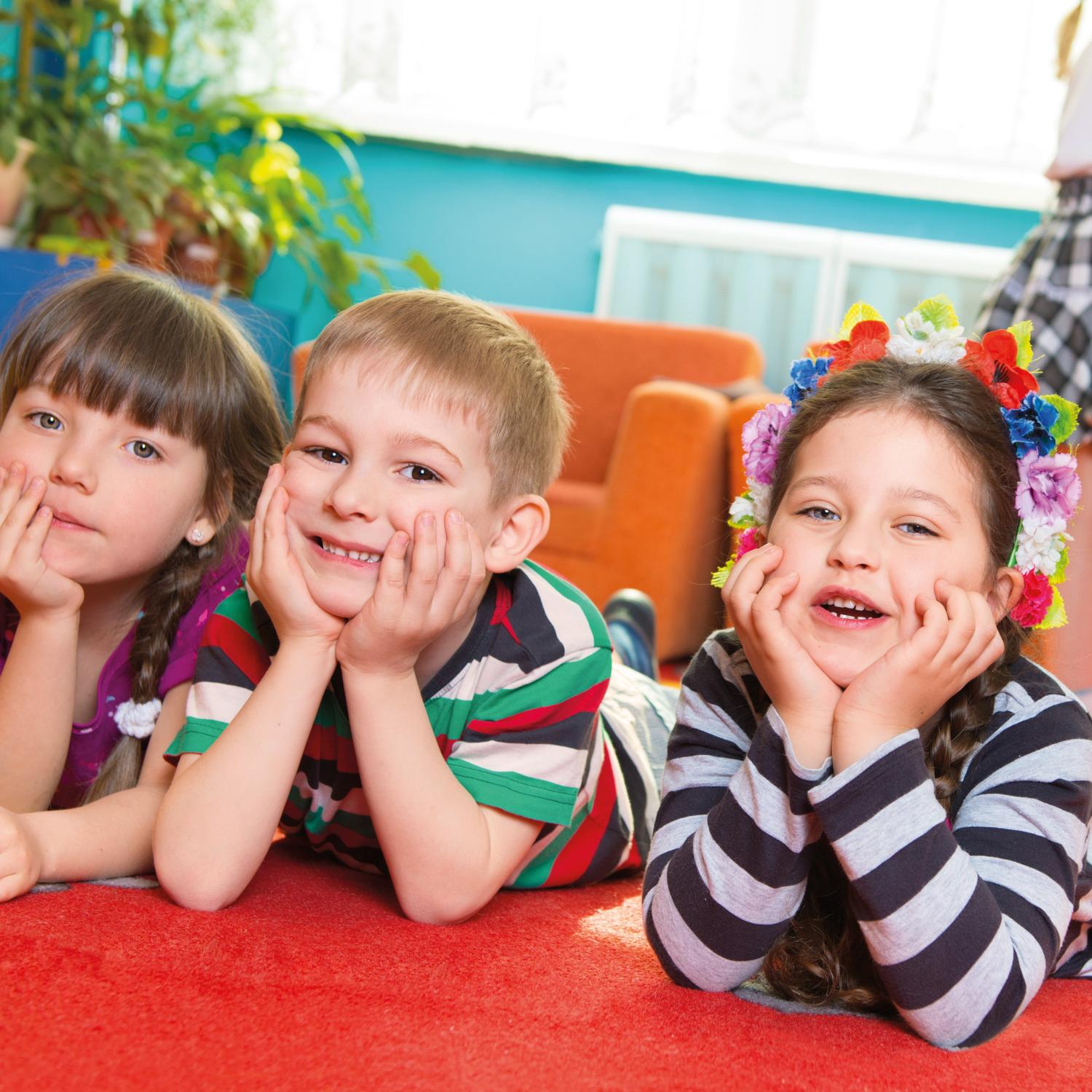Ein Junge und zwei Mädchen schauen im Kindergarten lachend in die Kamera. Sie liegen auf einer Spielmatte und haben Ihre Köpfe in die Hände gestützt