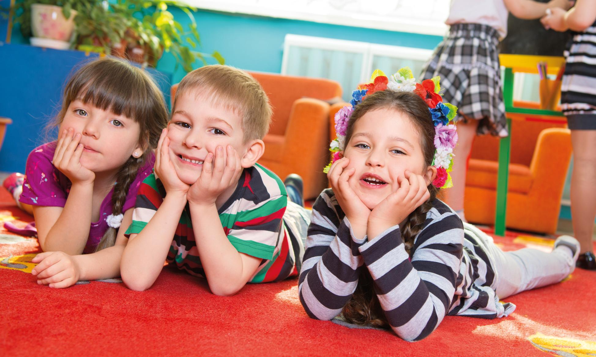 Ein Junge und zwei Mädchen schauen im Kindergarten lachend in die Kamera. Sie liegen auf einer Spielmatte und haben Ihre Köpfe in die Hände gestützt