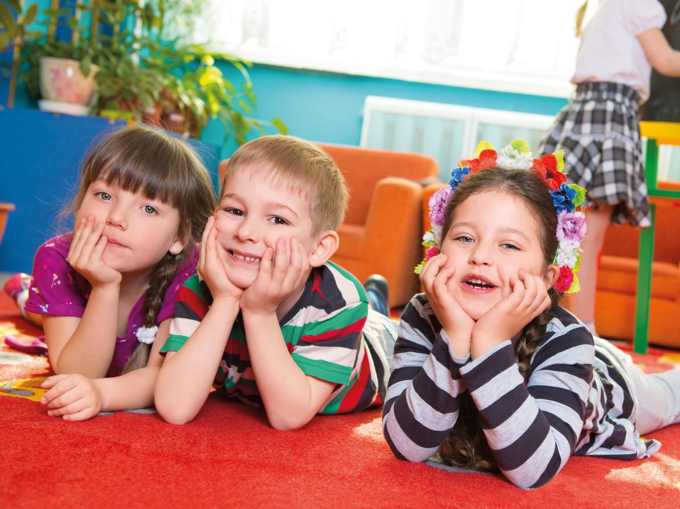 Ein Junge und zwei Mädchen schauen im Kindergarten lachend in die Kamera. Sie liegen auf einer Spielmatte und haben Ihre Köpfe in die Hände gestützt