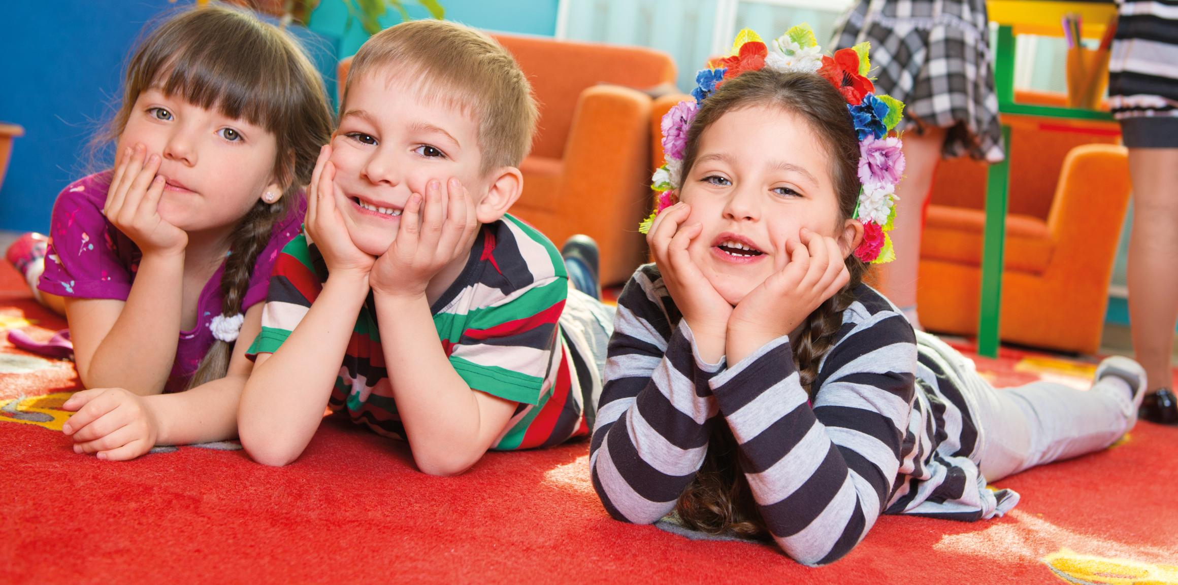 Ein Junge und zwei Mädchen schauen im Kindergarten lachend in die Kamera. Sie liegen auf einer Spielmatte und haben Ihre Köpfe in die Hände gestützt