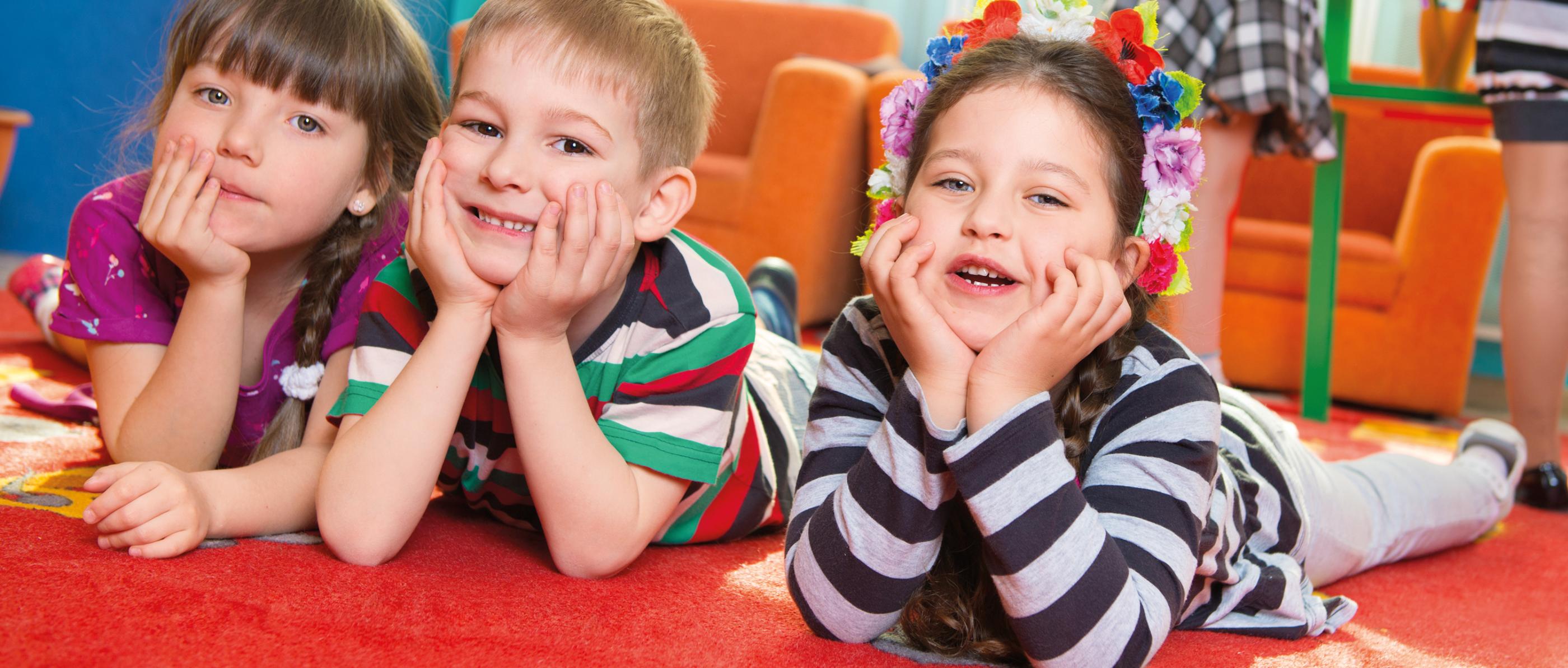 Ein Junge und zwei Mädchen schauen im Kindergarten lachend in die Kamera. Sie liegen auf einer Spielmatte und haben Ihre Köpfe in die Hände gestützt