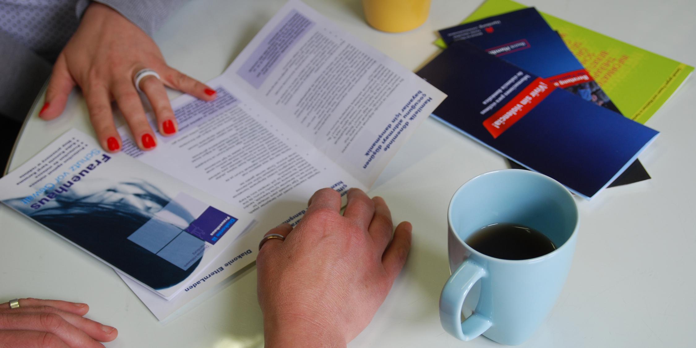 Zwei Personen sitzen an einem Tisch. Sie lesen Frauenhaus-Prospekte und trinken Tee.