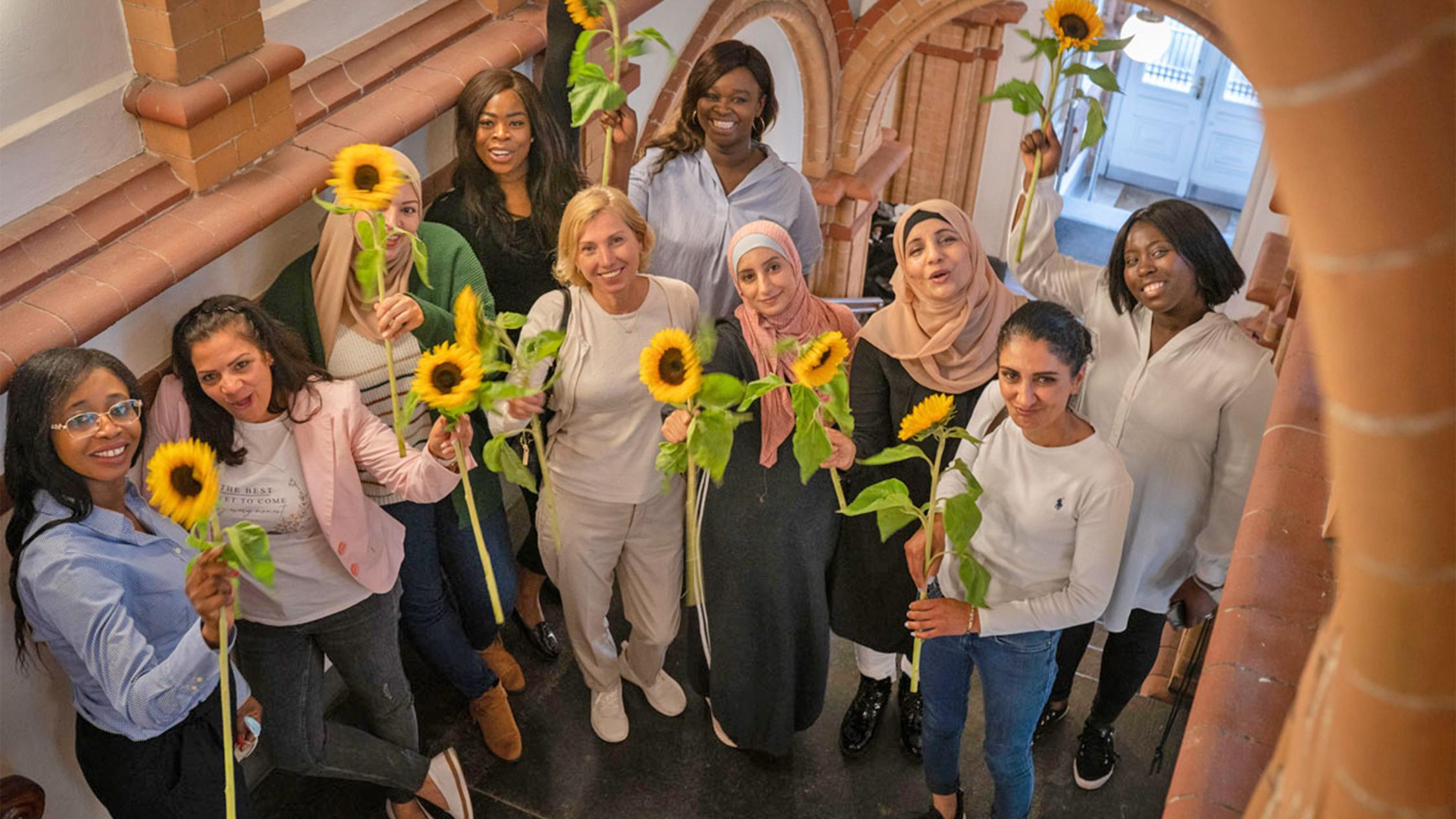 Mehrere Frauen unteschiedlicher Nationalitäten stehen in einem Treppenhaus und halten Sonnenblumen in der Hand.