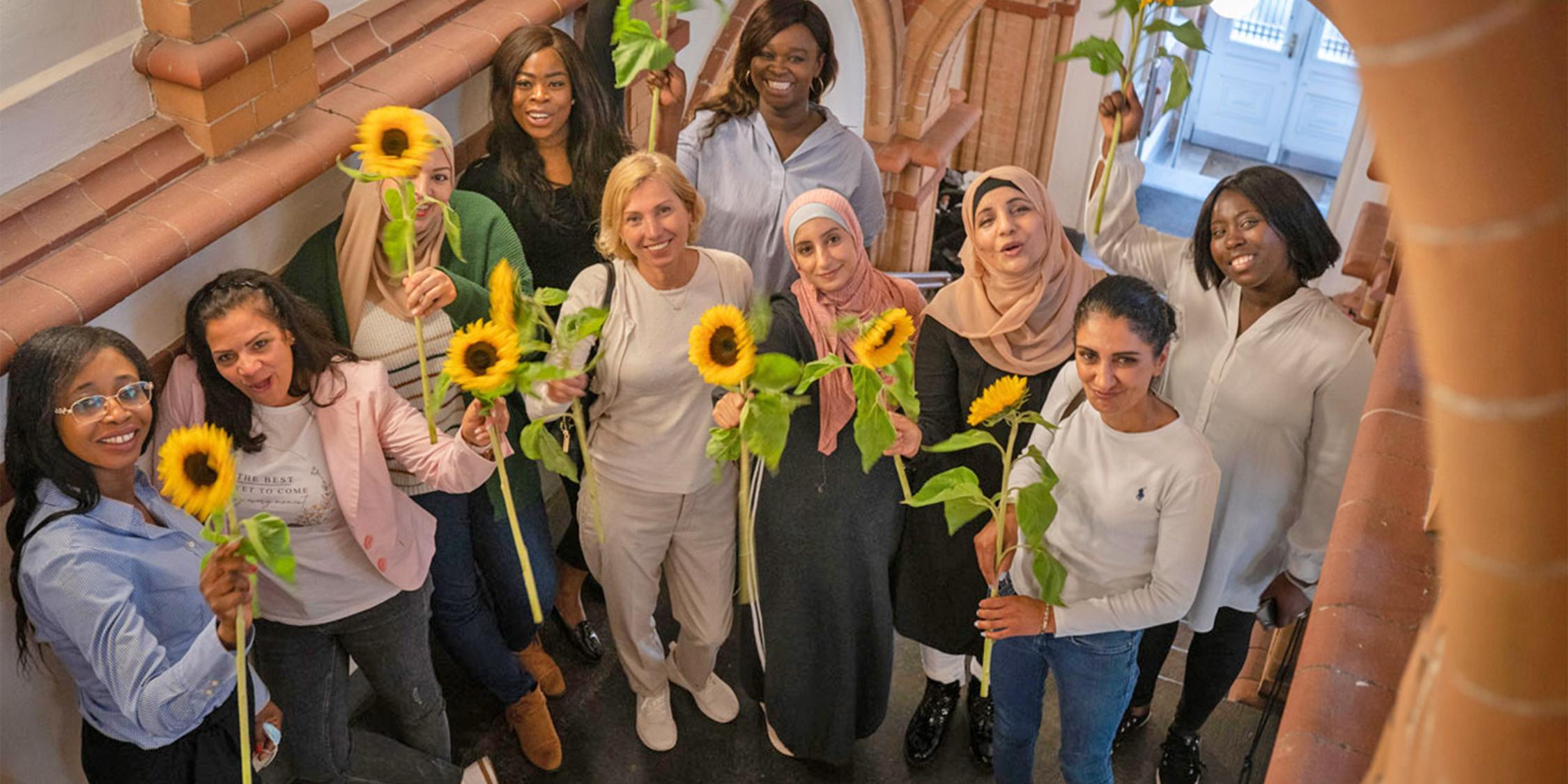 Mehrere Frauen unteschiedlicher Nationalitäten stehen in einem Treppenhaus und halten Sonnenblumen in der Hand.