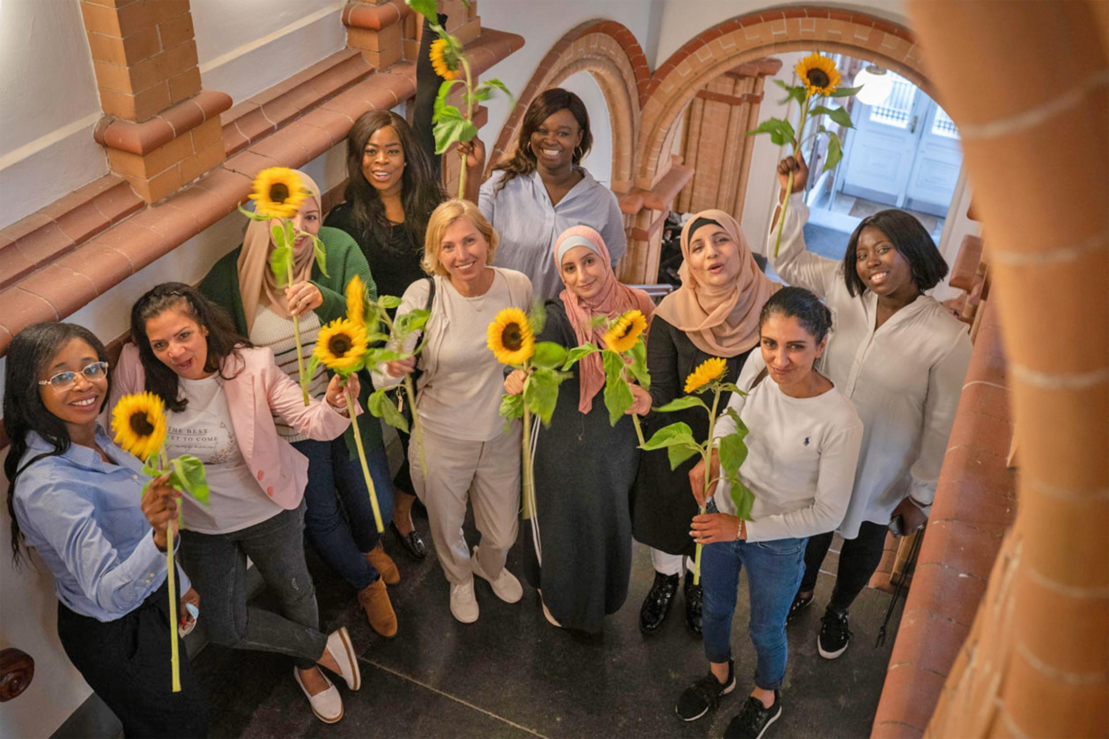 Mehrere Frauen unteschiedlicher Nationalitäten stehen in einem Treppenhaus und halten Sonnenblumen in der Hand.
