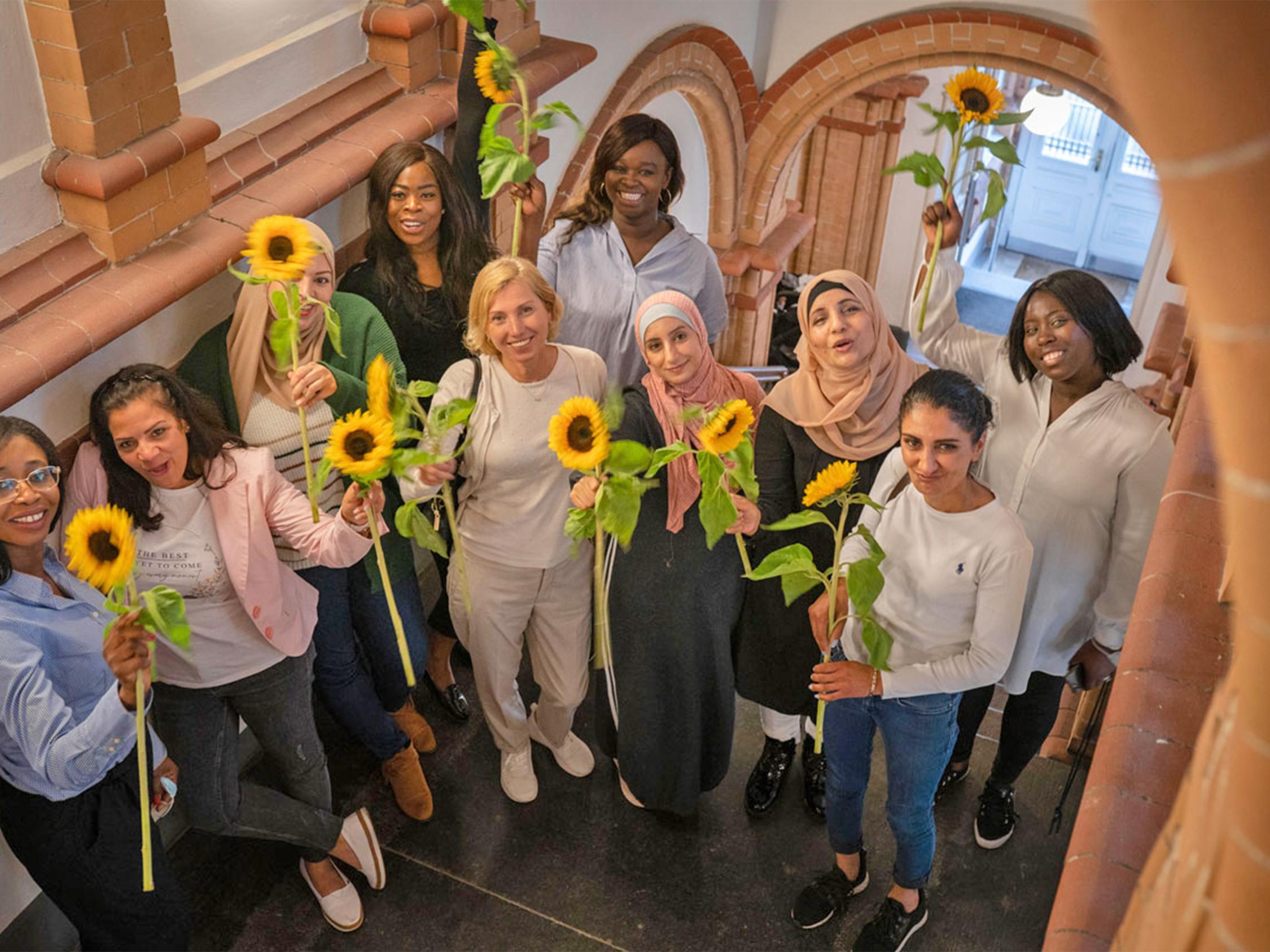 Mehrere Frauen unteschiedlicher Nationalitäten stehen in einem Treppenhaus und halten Sonnenblumen in der Hand.