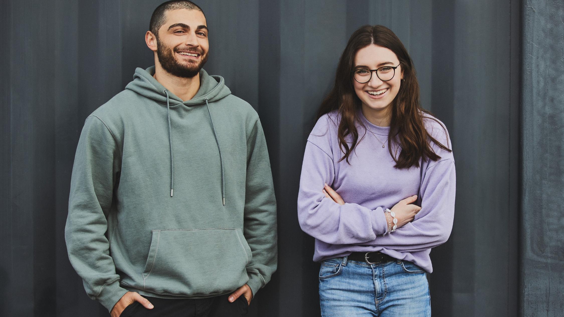 Ein junger Mann und eine junge Frau lehnen an einer Wand. Beide lachen.
