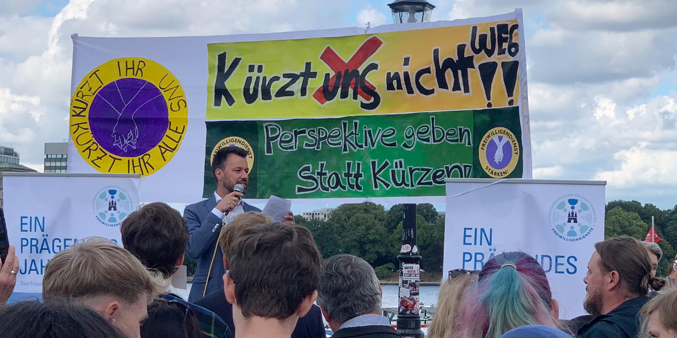 Ein Mann steht auf einer Bühne und spricht zu einer Gruppe von Menschen, von denen nur die Hinterköpfe zu sehen sind.