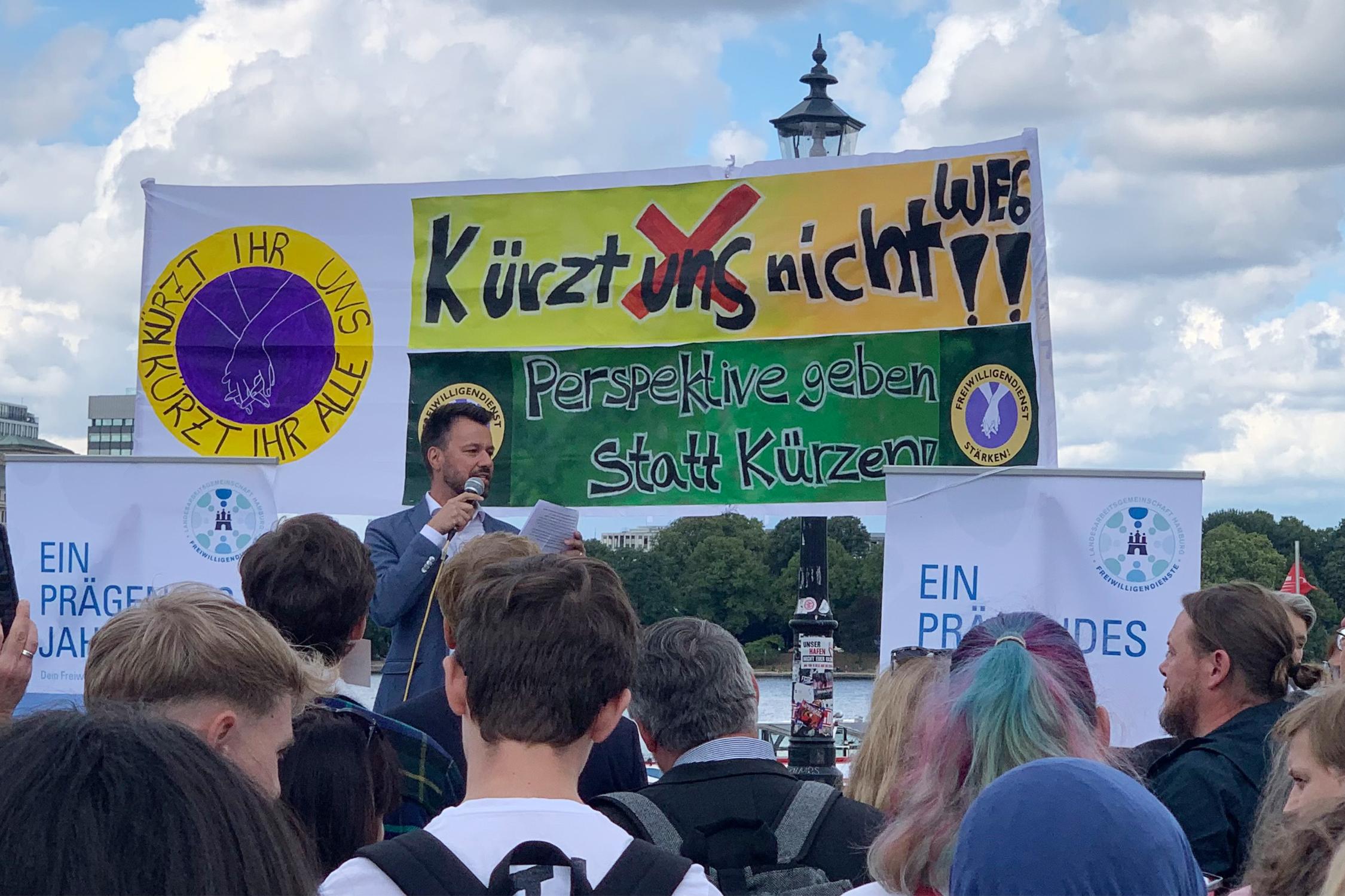 Ein Mann steht auf einer Bühne und spricht zu einer Gruppe von Menschen, von denen nur die Hinterköpfe zu sehen sind.