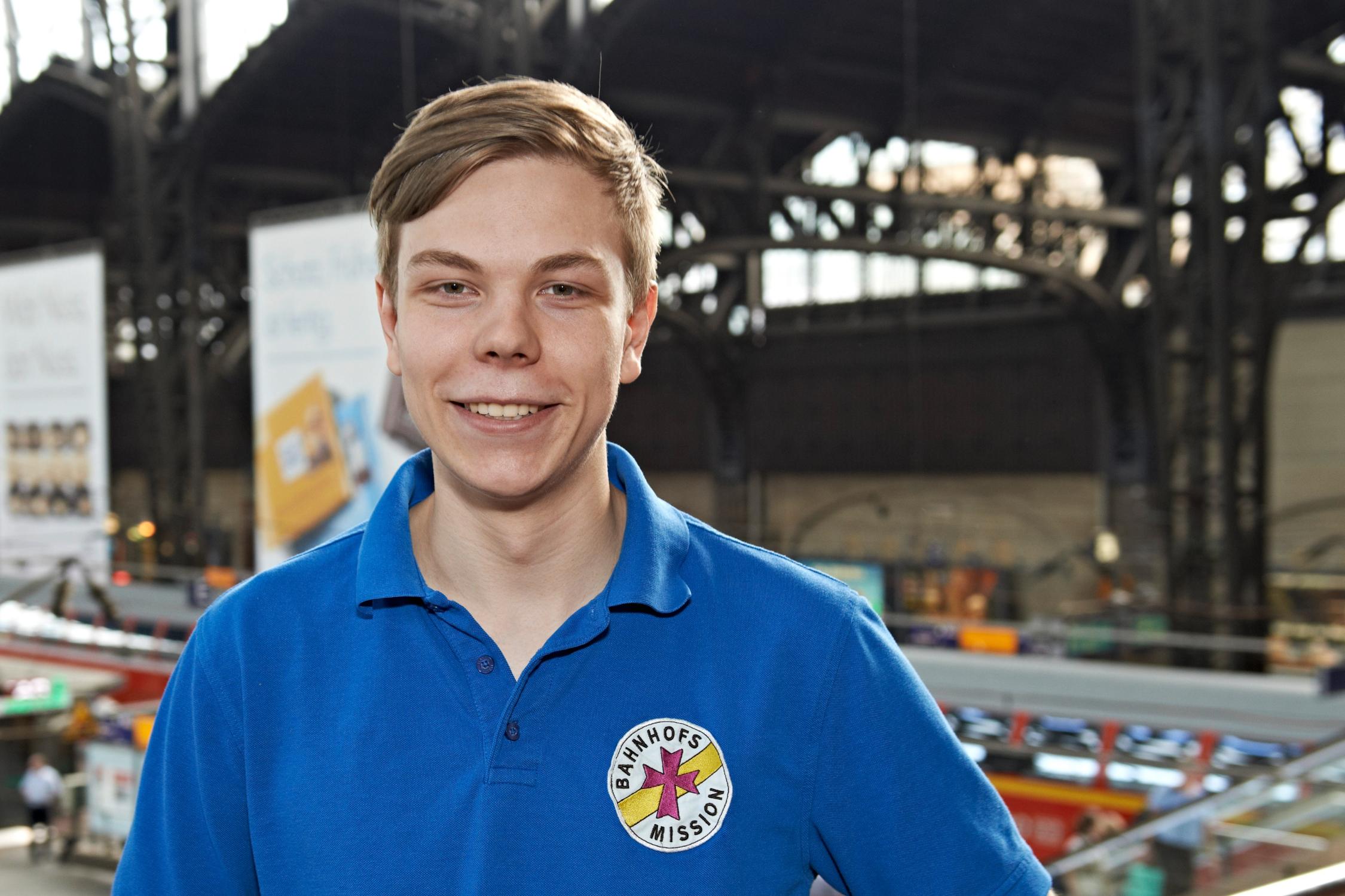 Ein junger Mann im T-Shirt der Bahnhofsmission steht im Hamburger Hauptbahnhof und lächelt in die Kamera.
