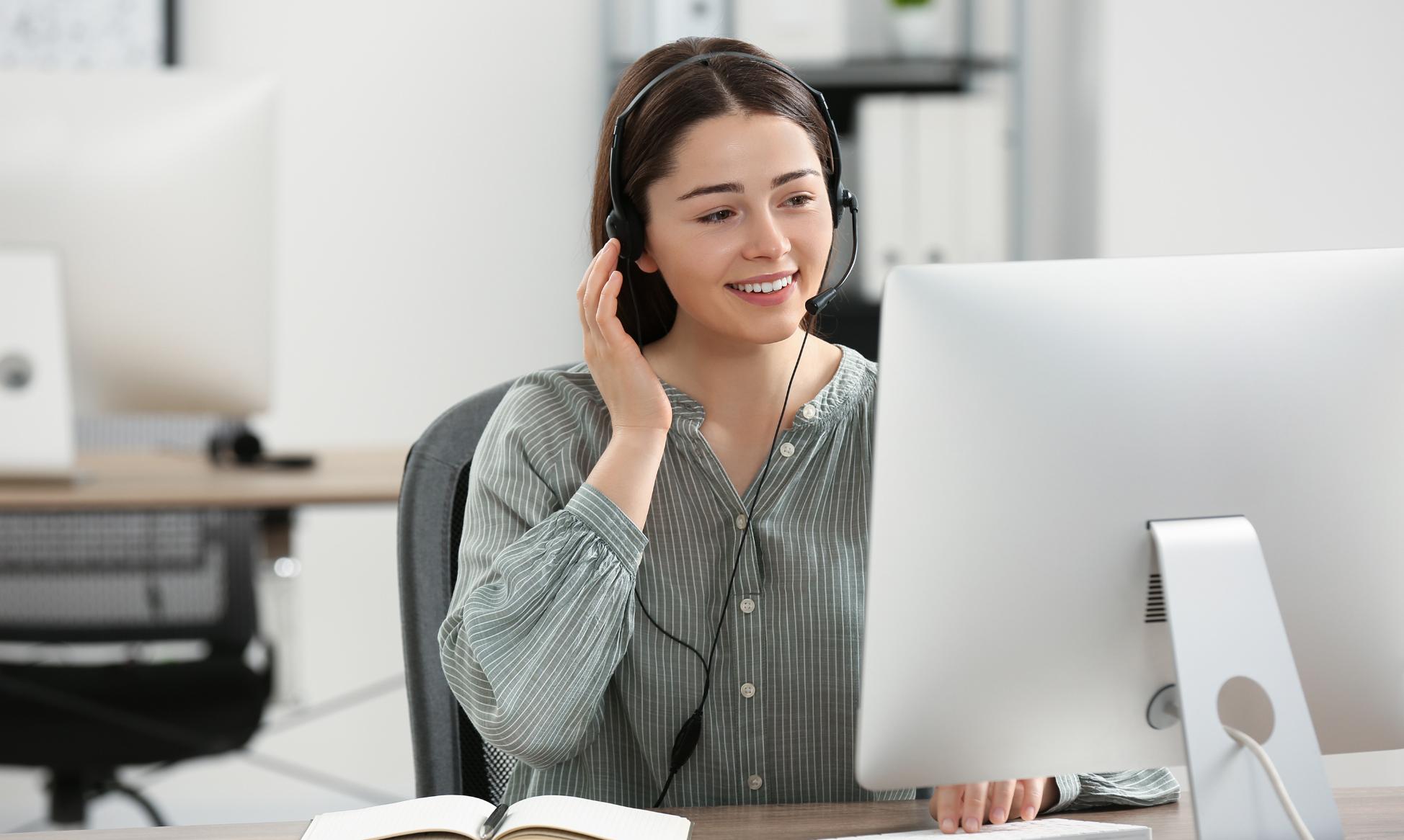 Eine lächelnde Frau mit Headset sitzt vor einem Computer und hält sich die Hand ans Ohr.
