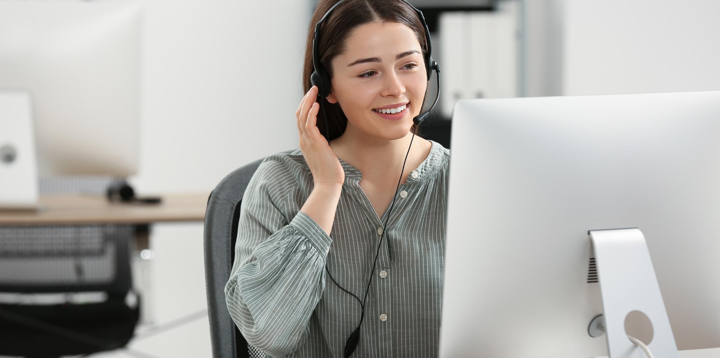 Eine lächelnde Frau mit Headset sitzt vor einem Computer und hält sich die Hand ans Ohr.
