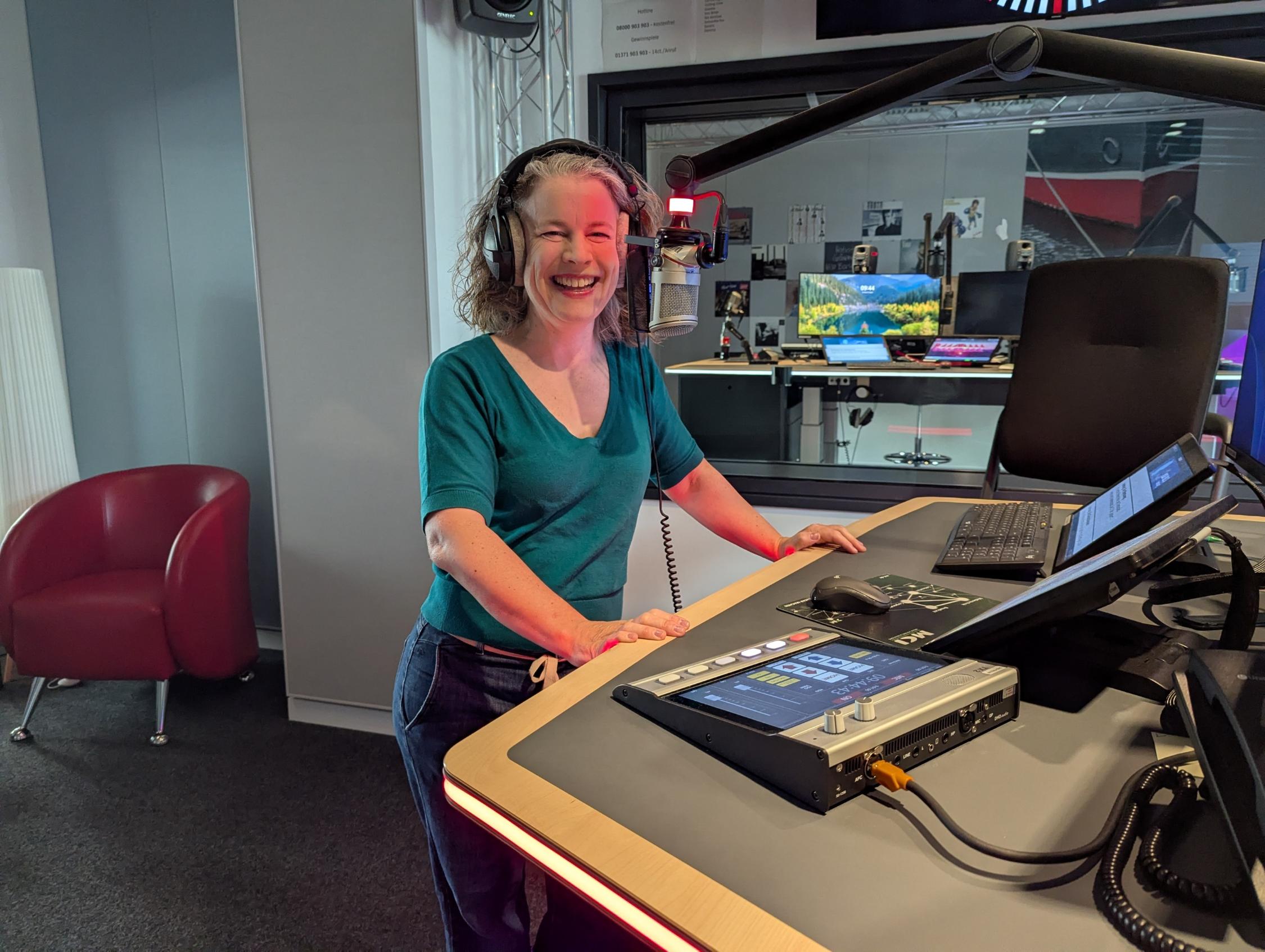 Lächelnde Frau mit Kopfhörern steht an einem Radiomischpult in einem Studio mit Mikrofon und Computerbildschirm