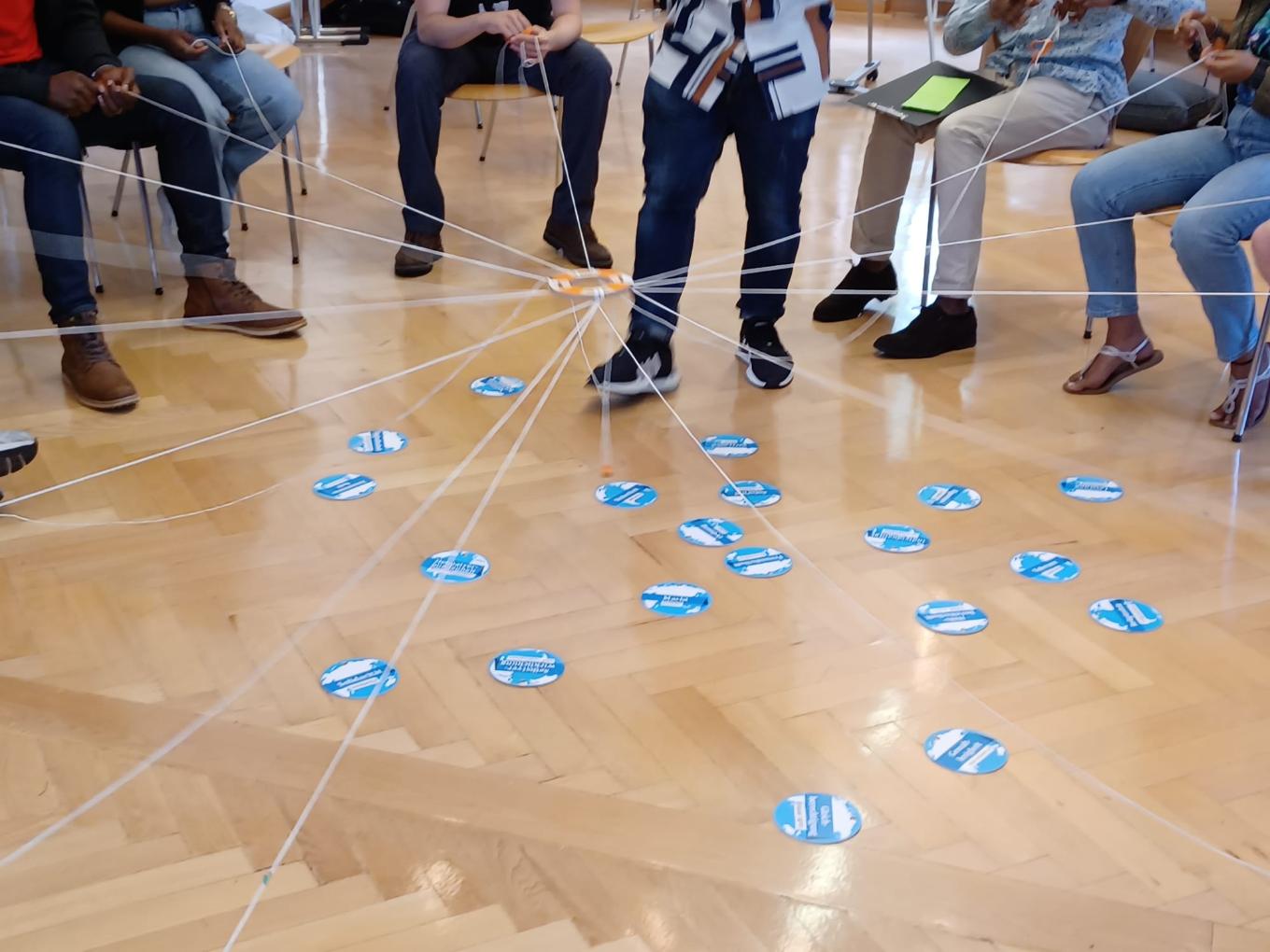 Personen, deren Köpfe man nicht sieht, sitzen im Stuhlkreis, halten Fäden in der Hand, die an einem zentralen Ring befestigt sind.