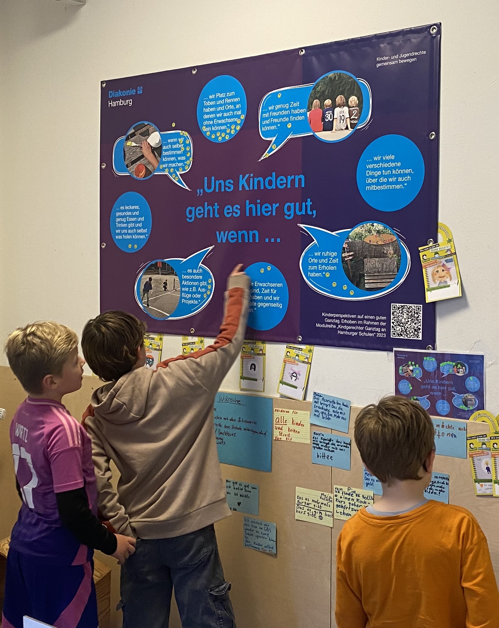 drei Kinder stehen vor einem Plakat der Diakonie zum Thema Kinderrechte und zeigen darauf.