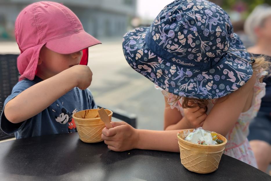 Zwei Kinder mit Sonnenhüten sitzen an einem Tisch und essen Eis aus Waffelbechern