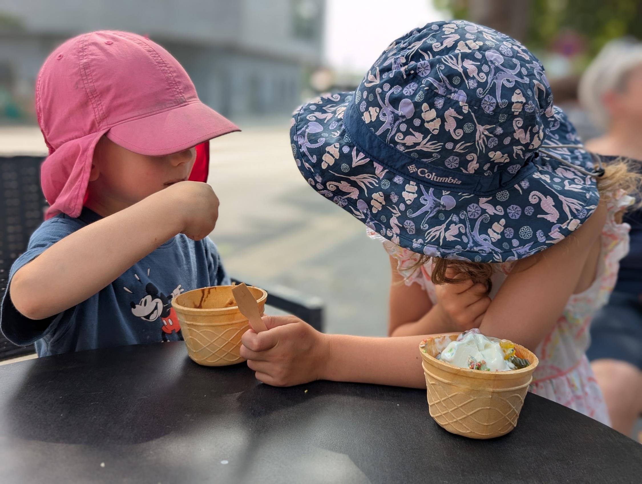 Zwei Kinder mit Sonnenhüten sitzen an einem Tisch und essen Eis aus Waffelbechern