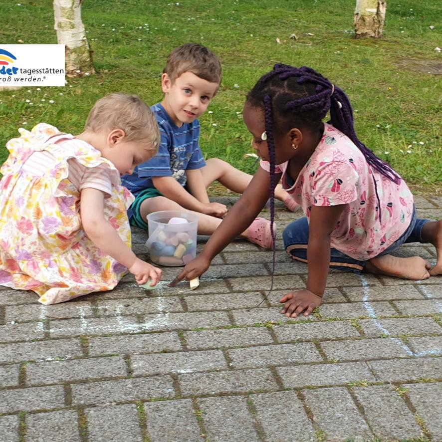 Drei Kinder sitzen auf einem gepflasterten Boden und spielen mit kleinen Gegenständen