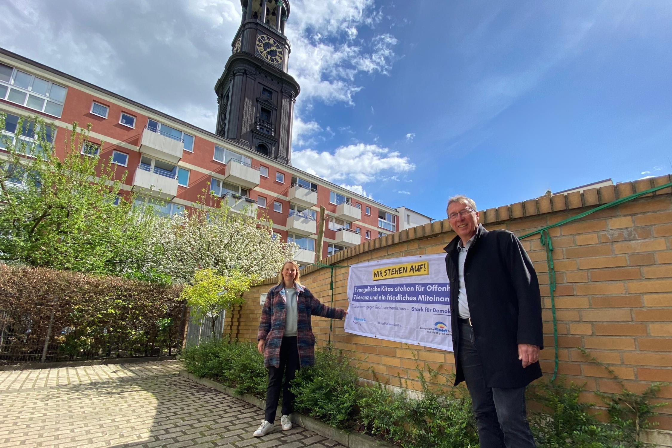 Ein Mann und eine Frau stehen lächelnd vor einer Mauer, an der ein Plakat hängt. Die Frau zeigt darauf.