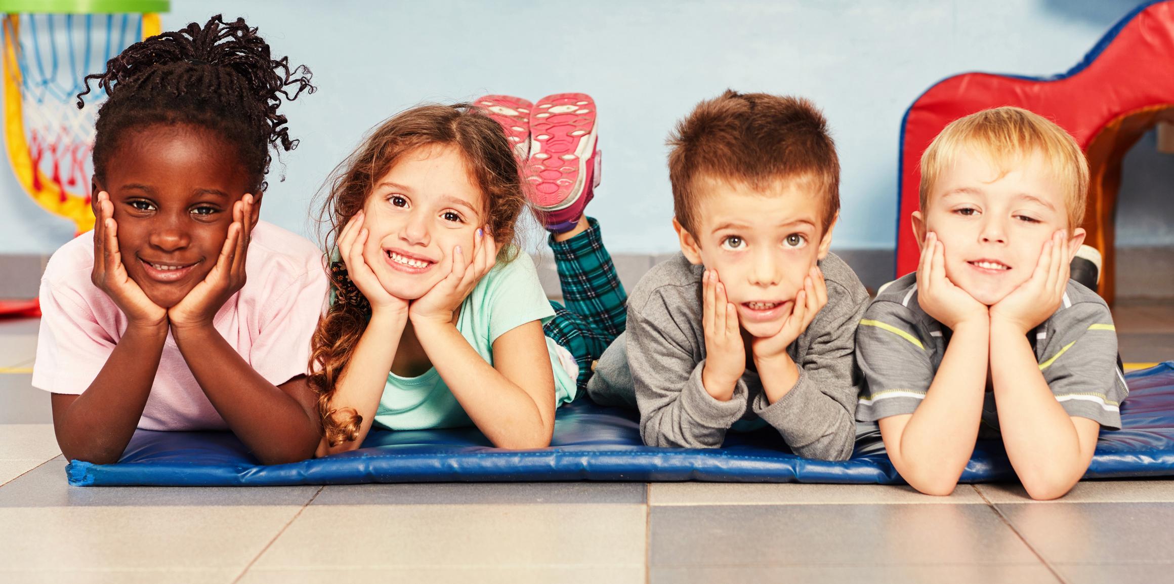 Vier Kinder liegen bäuchlings nebeneinander auf einer blauen Matte, den Kopf in die Hände gestützt, und lächeln in die Kamera.