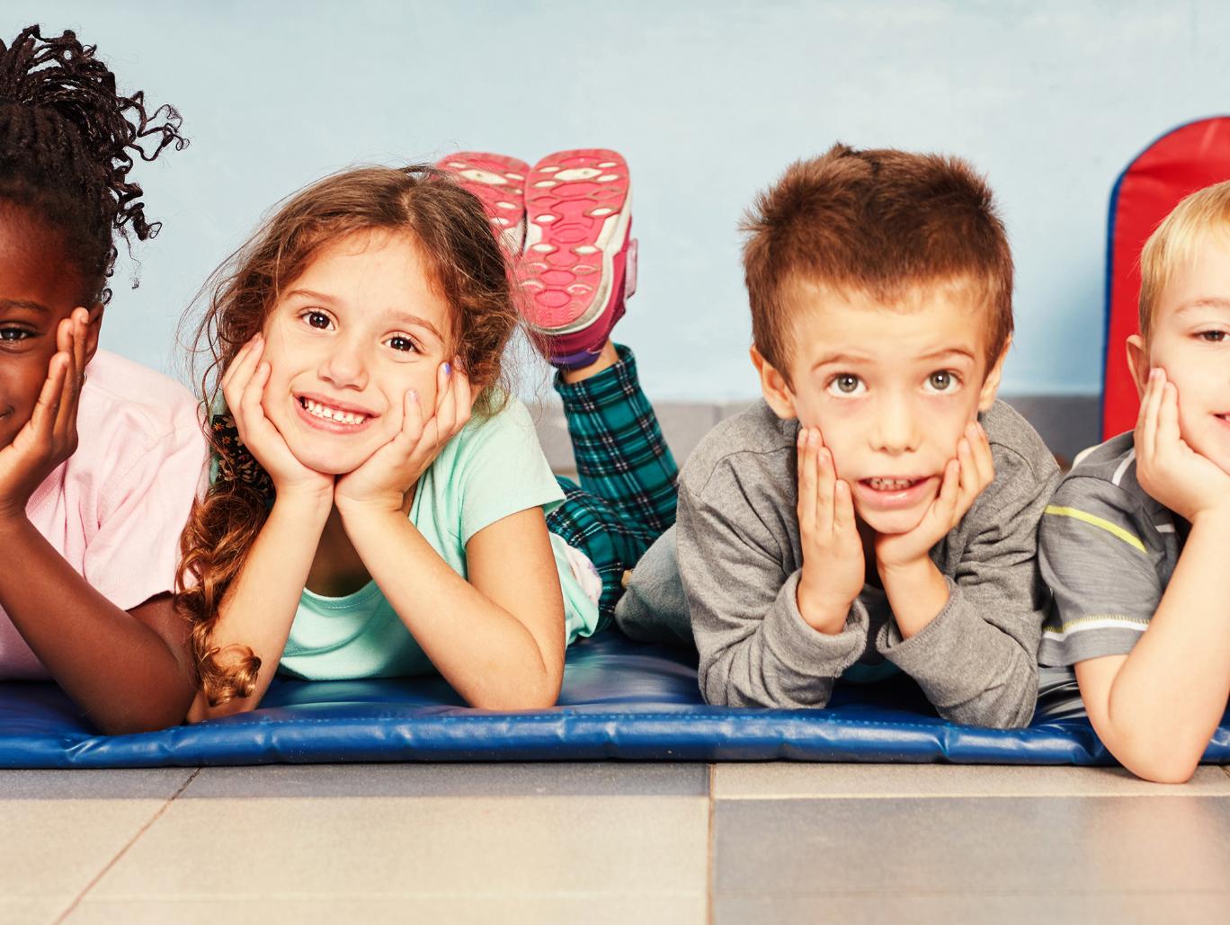 Vier Kinder liegen bäuchlings nebeneinander auf einer blauen Matte, den Kopf in die Hände gestützt, und lächeln in die Kamera.
