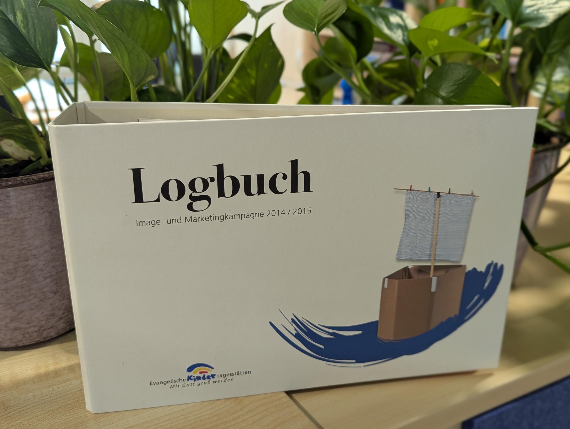 Logbuch mit Titel und Illustration eines Papierschiffs auf einem Holztisch vor grünen Pflanzen