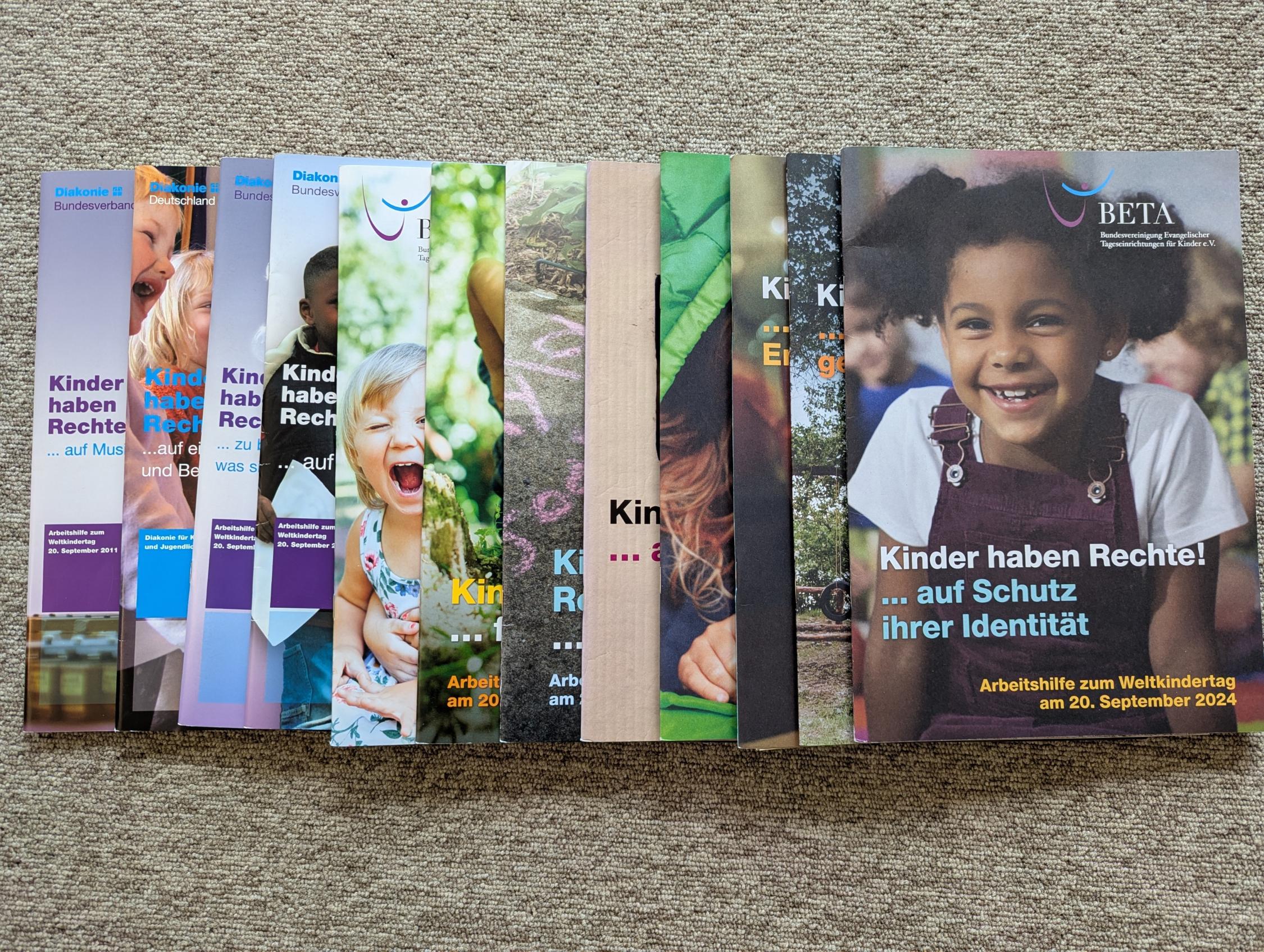 Mehrere Broschüren mit Bildern von Kindern und Texten wie 'Kinder haben Rechte' liegen fächerförmig auf einem roten Stoff.