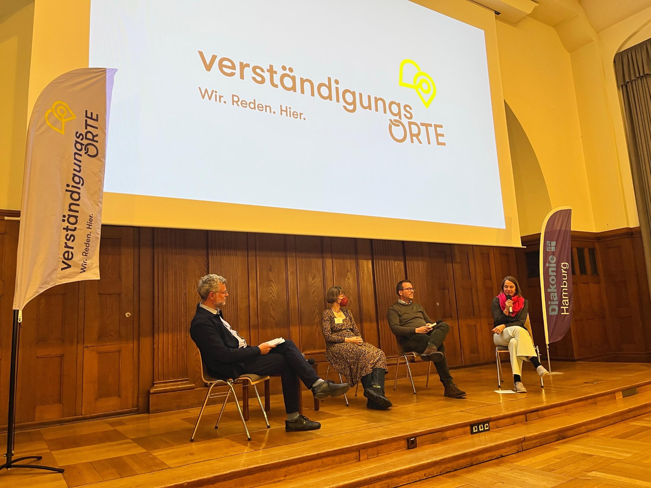 Vier Menschen sitzen auf einem Podium und unterhalten sich. Zu sehen sind zwei Männer und zwei Frauen.