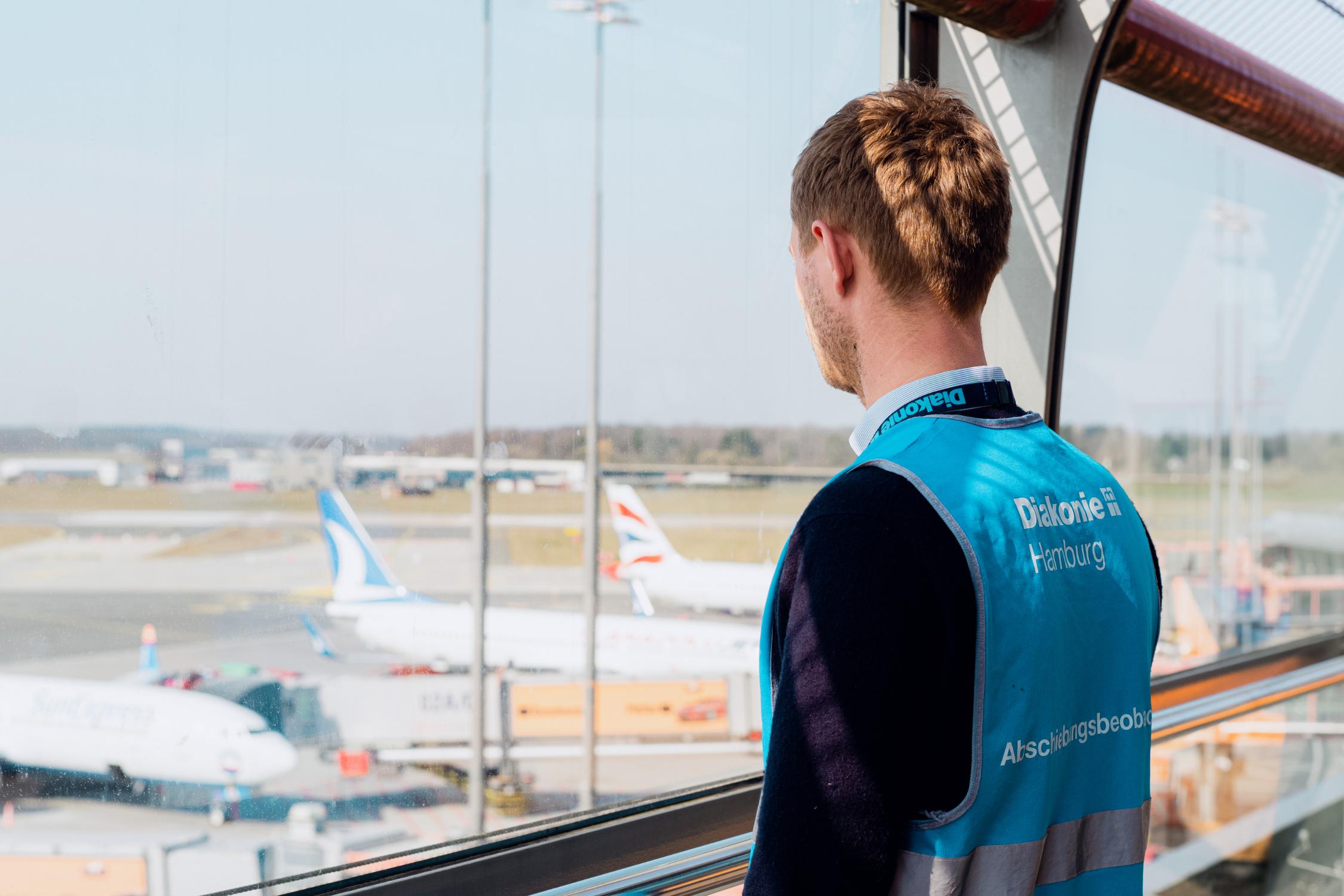 Ein Mann mit blauer Weste der Diakonie schaut aus dem Fenster herunter auf eine Rollbahn am Flughafen.