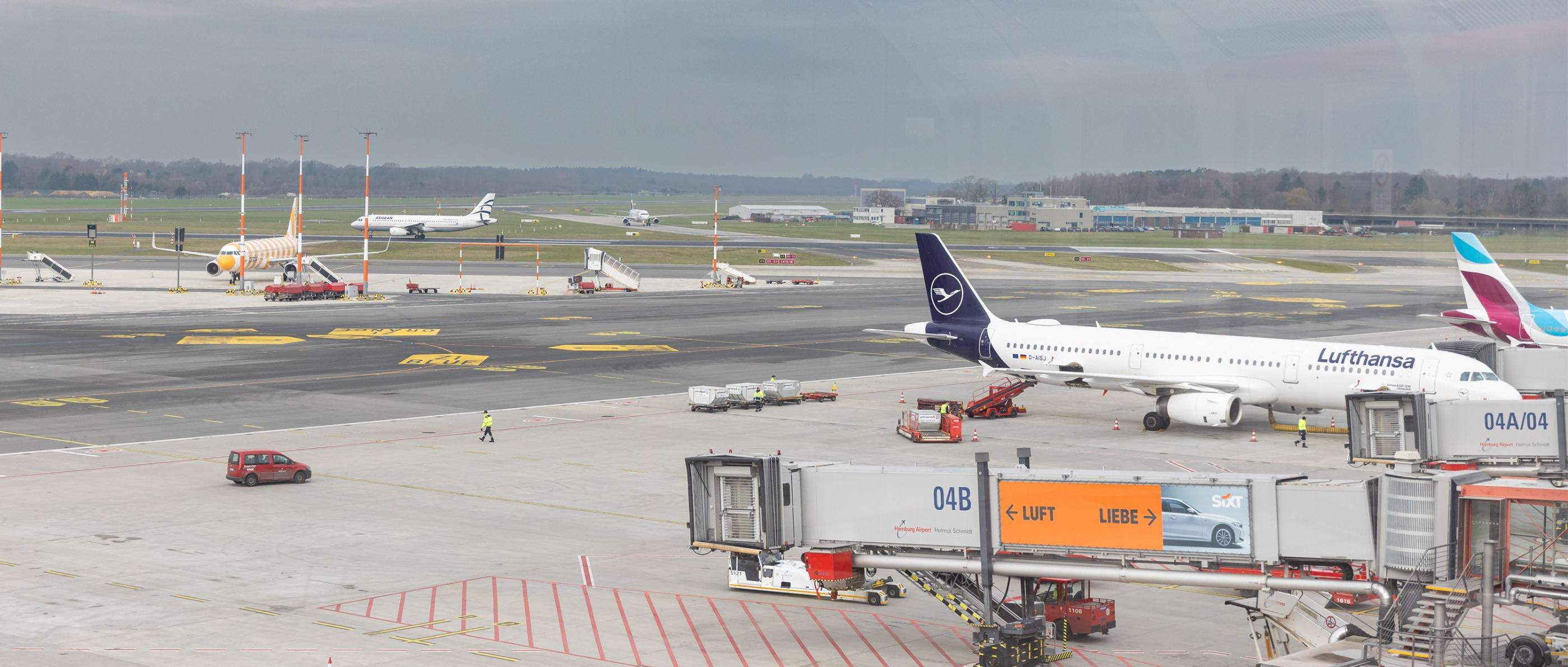 Durch eine Scheibe blickt man auf die Rollbahn eines Flughafens. Überall stehen Flugzeuge, Autos und Flugzeugtreppen.