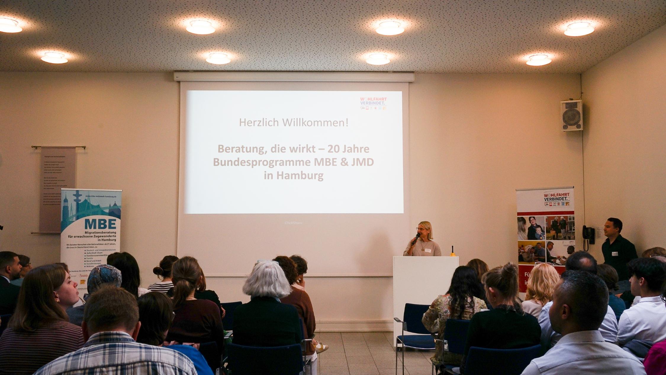 Menschen sitzen in einem Veranstaltungsraum. Eine Frau steht vorne und hält einen Vortrag.