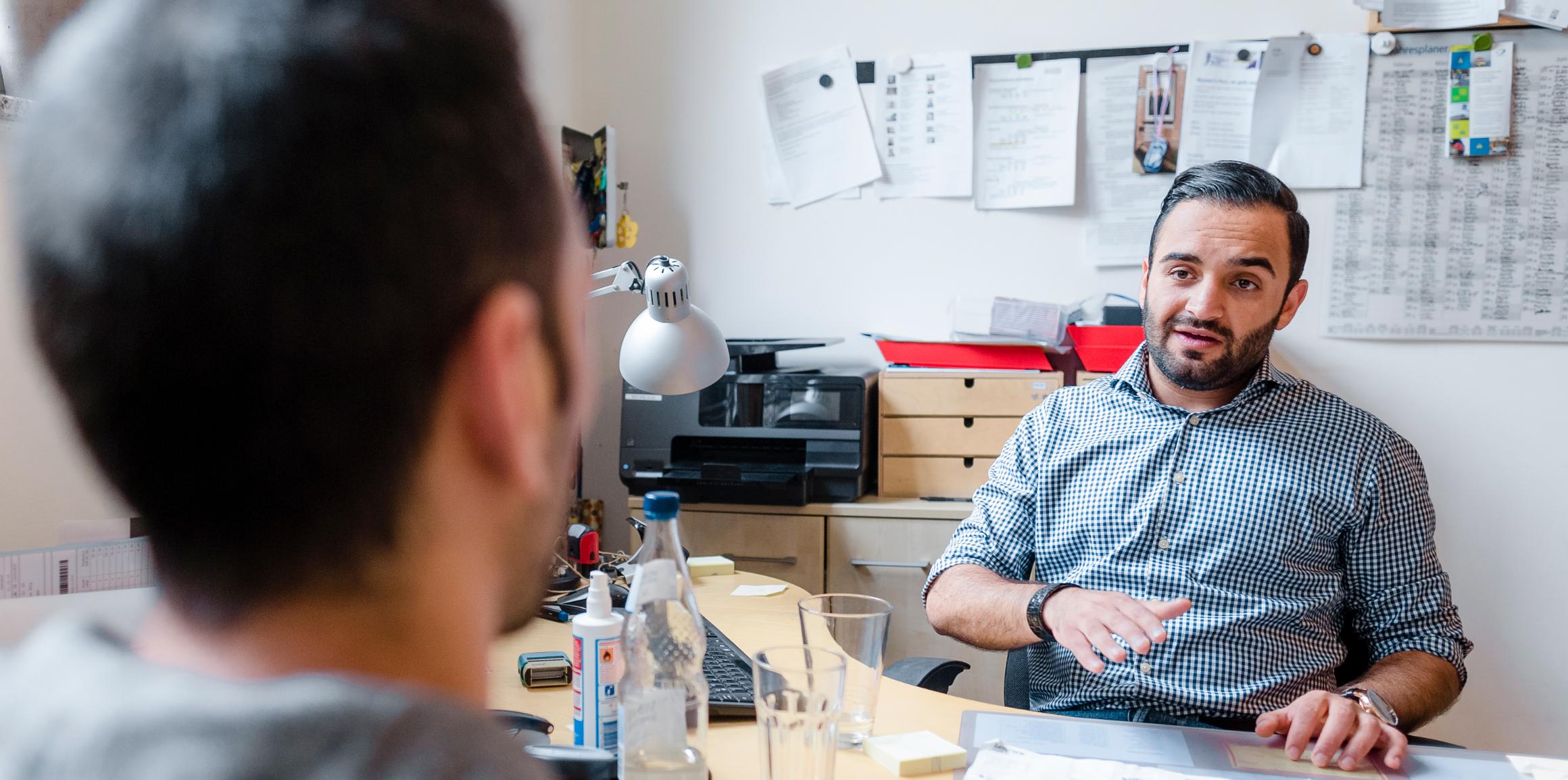 Ein Mann sitzt an einem Schreibtisch in einem Büro und spricht mit einem anderen Mann, der ihm gegenüber sitzt.