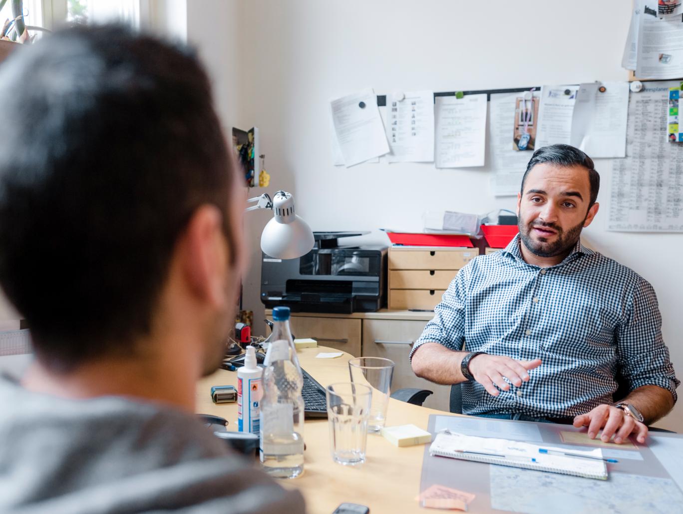 Ein Mann sitzt an einem Schreibtisch in einem Büro und spricht mit einem anderen Mann, der ihm gegenüber sitzt.