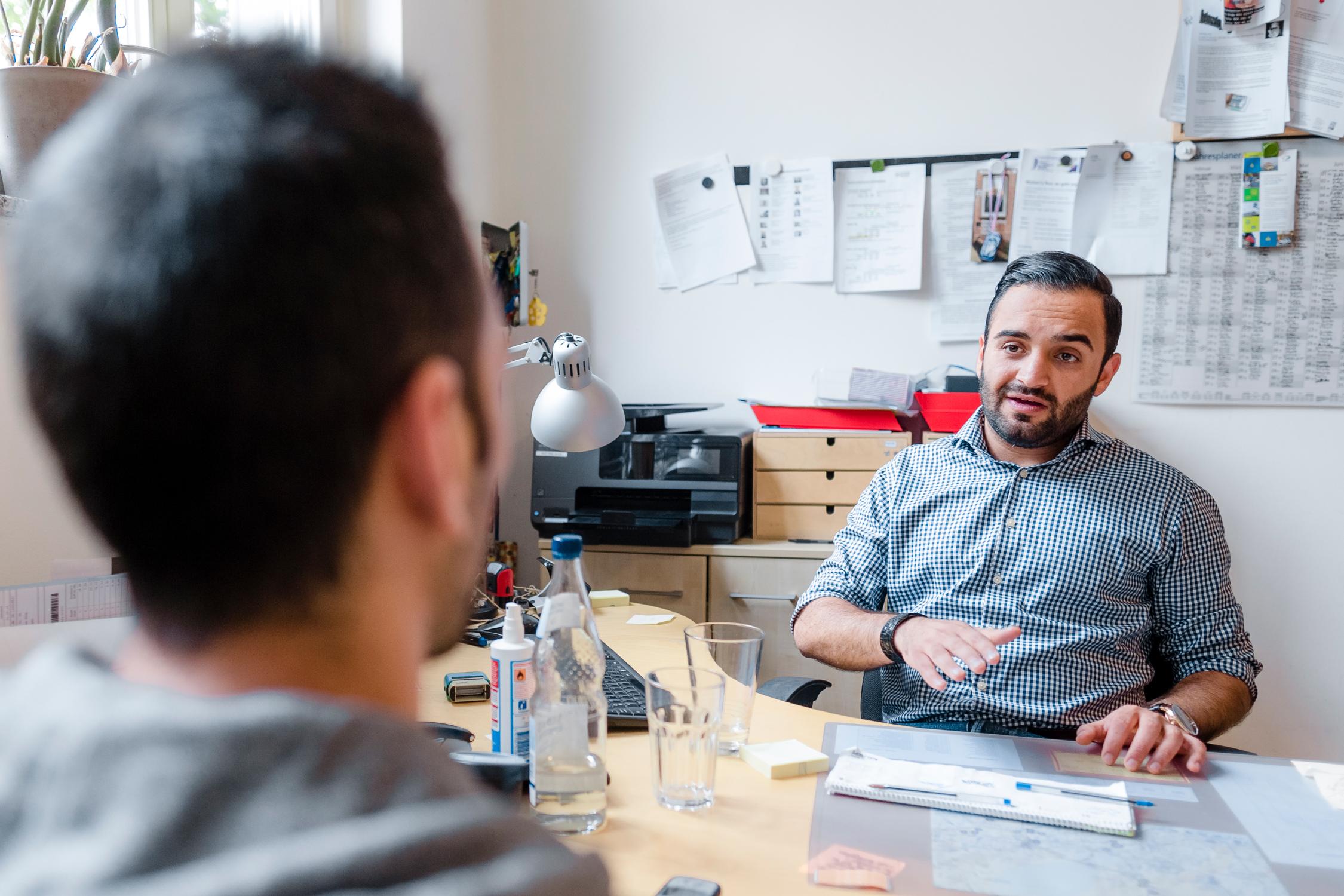 Ein Mann sitzt an einem Schreibtisch in einem Büro und spricht mit einem anderen Mann, der ihm gegenüber sitzt.
