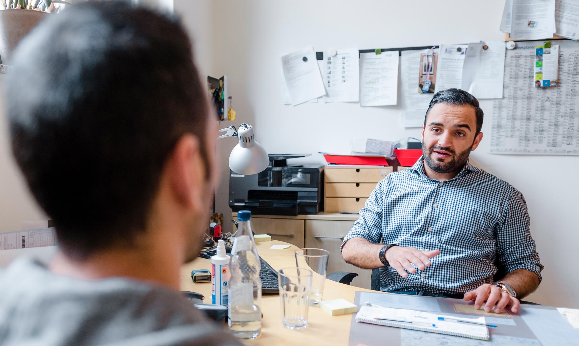 Ein Mann sitzt an einem Schreibtisch in einem Büro und spricht mit einem anderen Mann, der ihm gegenüber sitzt.