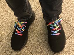 Auf einem Gehweg stecken zwei Füße in einem Paar schwarzer Schuhe mit bunten Schnürsenkeln.