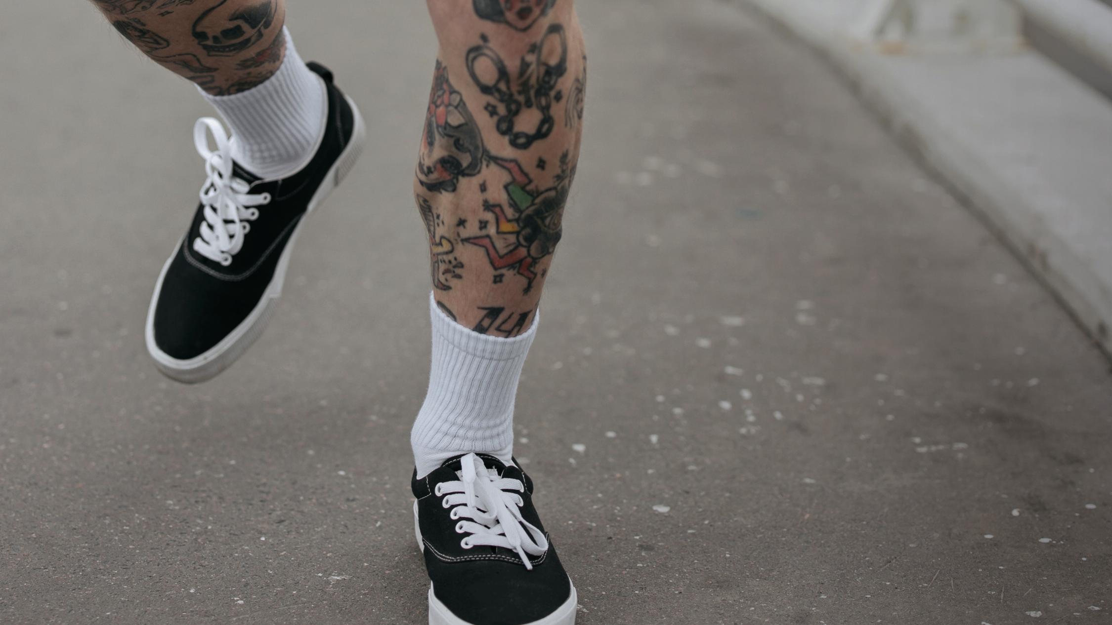 Tattoowierte Beine in einem Paar schwarz-weißer Sneaker mit hohen langen Socken, die über Asphalt laufen.