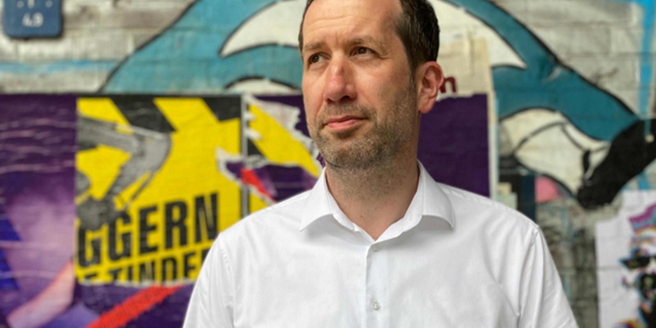 Ein Mann im Portrait steht vor einer Graffiti-Mauer und schaut schräg nach links oben.